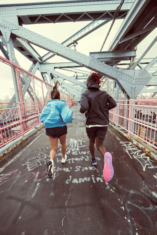 Two people jogging on a bridge with metal supports and pink railings, graffiti on the ground, wearing running shoes and active wear.