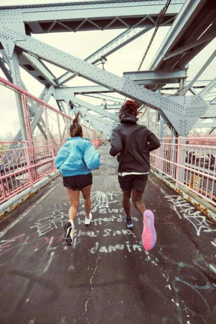 Two people jogging on a bridge with metal supports and pink railings, graffiti on the ground, wearing running shoes and active wear.