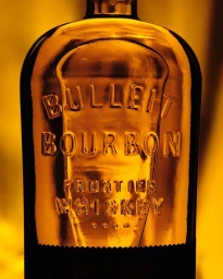 Close-up view of a glass bottle of Bulleit Bourbon whiskey lit with warm amber backlight, highlighting embossed lettering.