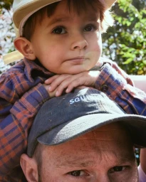 A young child sitting on a man's shoulders, wearing a plaid shirt and light cap, photographed in Los Angeles by Brian Kelley.