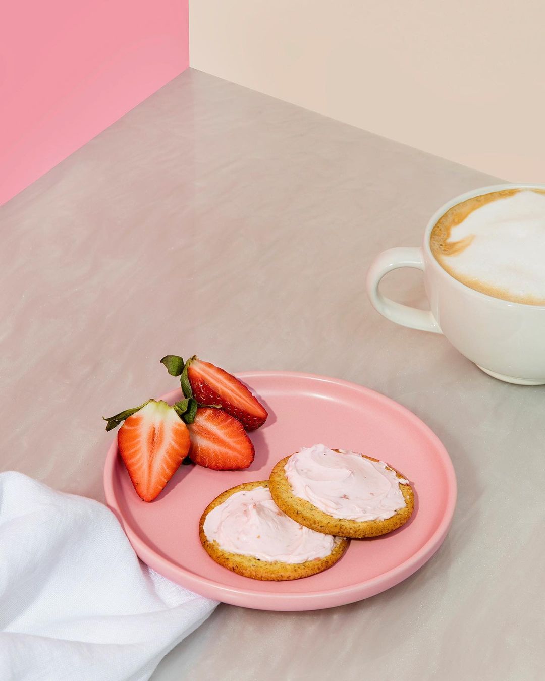 Crackers with pink topping and sliced strawberries displayed with a mug of coffee on a neutral-toned table against a two-tone backdrop.