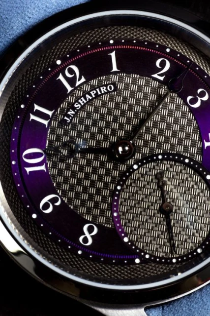 Close-up studio shot of a sophisticated watch with black hands, purple accents, and a textured metal face under bright lighting.