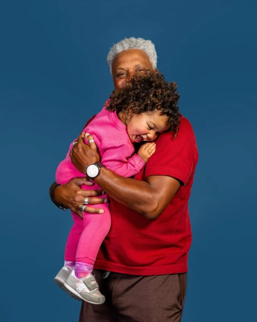 Elderly African-American man holds a toddler girl in a pink outfit against a dark blue background, creating a heartwarming intergenerational portrait.