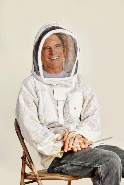 A smiling adult male beekeeper wearing a protective hooded suit sits on a wooden chair in a light studio setting. He is holding a tool.