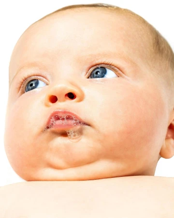 Close-up portrait of a baby with bright blue eyes looking upward with small bubbles around the lips against a clean white background.