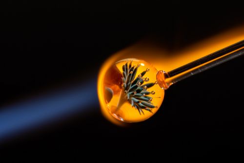 Close-up of a glassblowing process with molten glass on the end of a rod, illuminated by the bright yellow-orange flame of a torch against a black background.