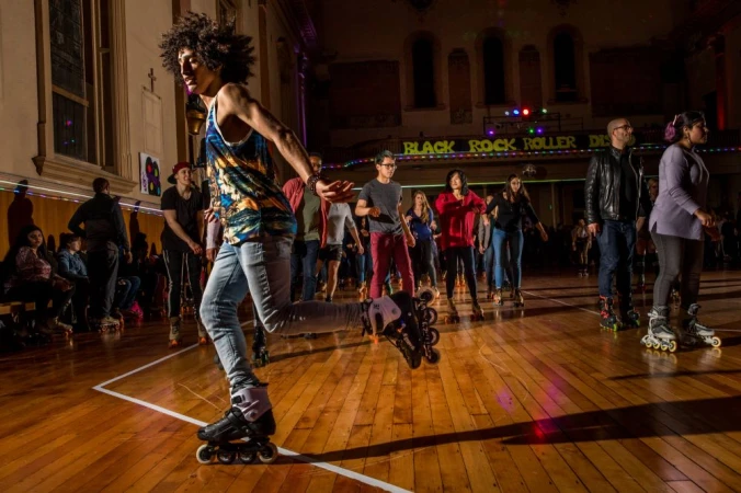 A roller skater with dark curly hair is skating indoors. The background shows a large group of people roller skating in a public place.