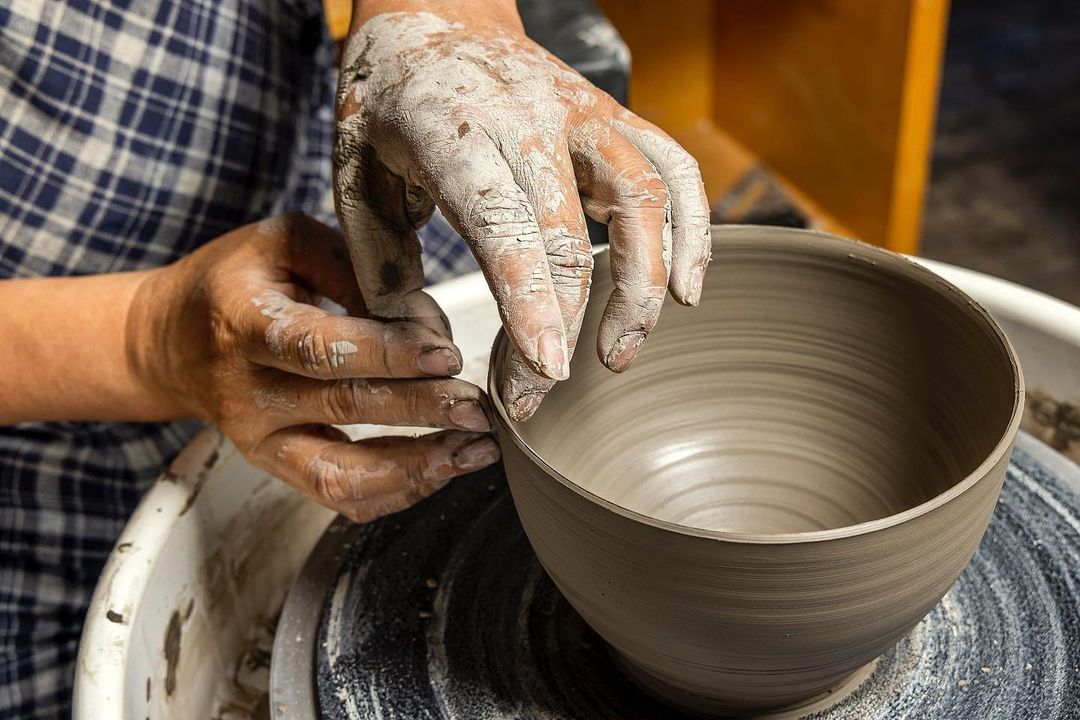 Close up on human hands forming a ceramic bowl on a pottery wheel. The potter is wearing a blue plaid shirt.