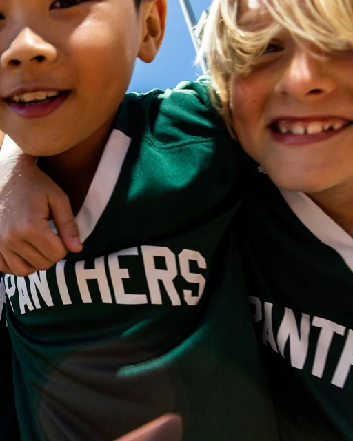 Close-up of two smiling boys wearing green and grey sports jerseys with 'Panthers' printed on them, outdoors on a bright day.