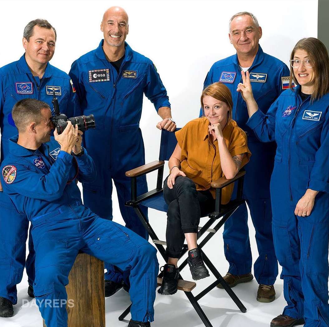Group photo featuring six individuals in a studio with a white background, some wearing blue jumpsuits and one seated in a director's chair.