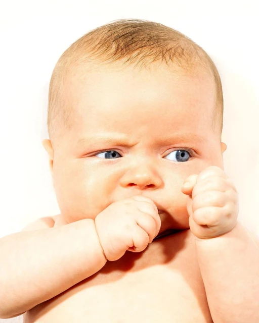 Infant with furrowed brow holding hands to mouth in front of a bright background, exhibiting a displeased expression, head and shoulders.