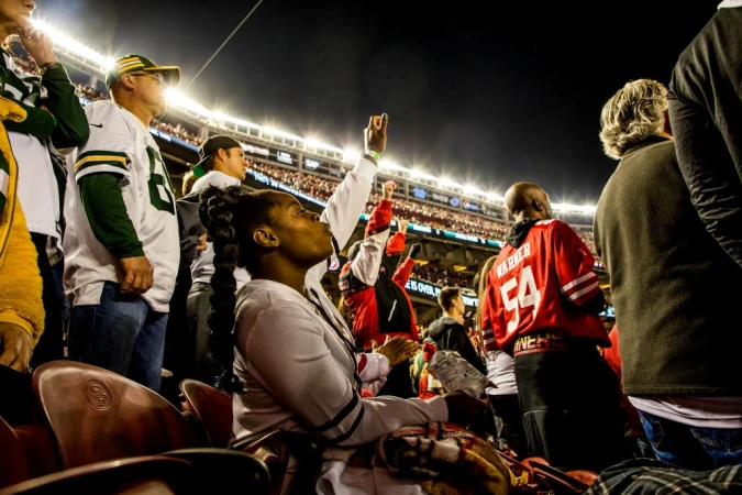 Fans in jerseys watch a football game at night in an outdoor stadium with bright lights, and dark sky behind the crowd.