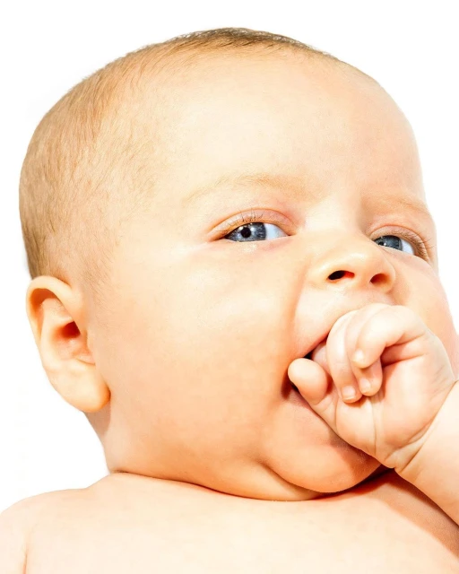 Close-up portrait of a baby with fair skin and blue eyes, sucking their thumb against a bright white background in a studio setting.