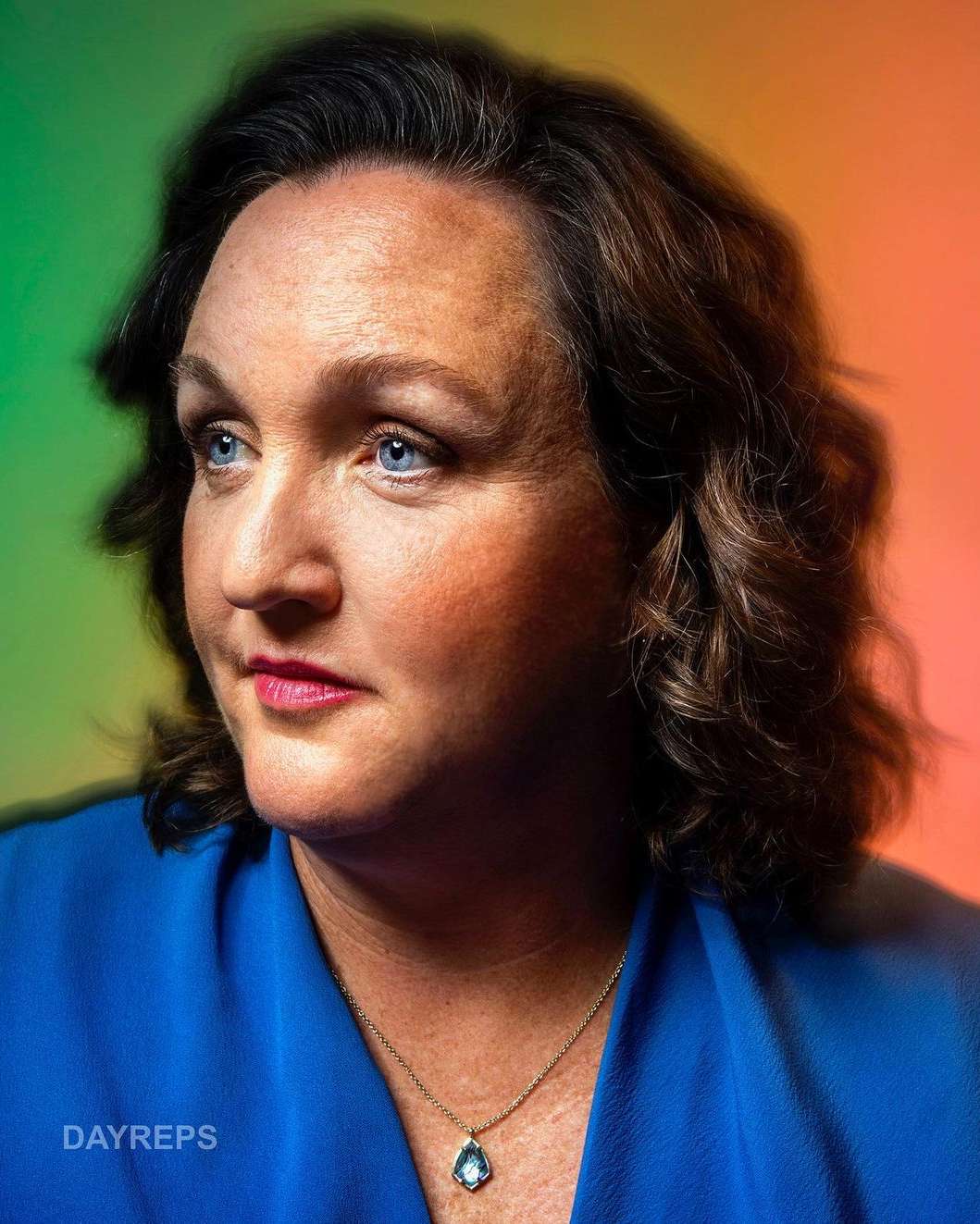 Portrait of an adult woman with blue eyes and wavy brown hair, wearing a blue blouse and a necklace with a blue pendant.