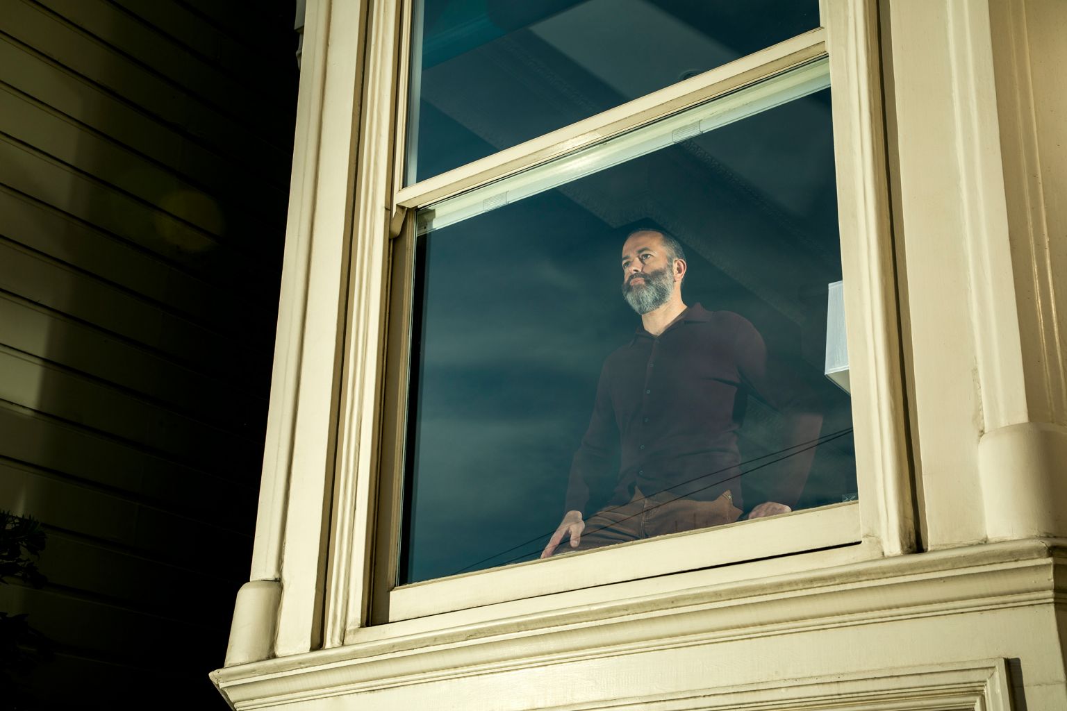 A bearded man in a burgundy shirt and brown pants kneels inside by a large window, looking out into the soft, cloudy night.