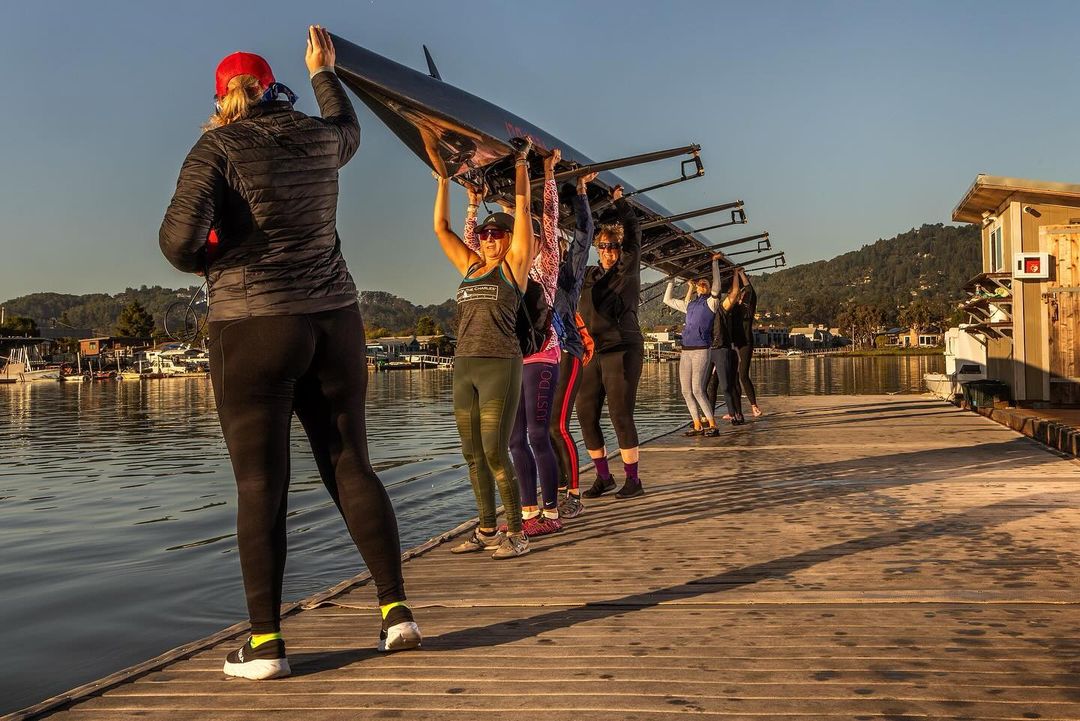 Eight women in athletic wear carry a racing shell on a wooden dock next to water. The sky is clear and the sun is shining.