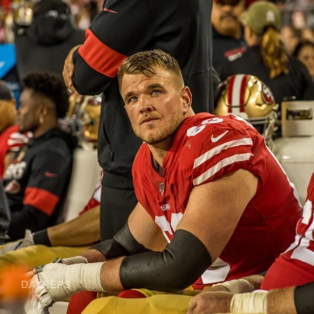 American football player wearing a red and white jersey with bandages, sitting on a bench during a game with crowd in background.