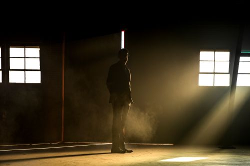 Silhouette of a man standing indoors with natural light streaming through windows in a dark and sparse environment, creating a moody atmosphere.