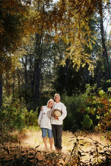 A couple stands in a sunlit forest. The woman wears a white jacket and blue shorts. The man wears a white jacket, dark pants and holds a hat.