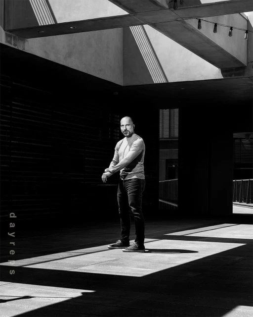 Black and white portrait of a bald adult male standing outdoors in casual clothing, with overhead architectural beams and shadows.