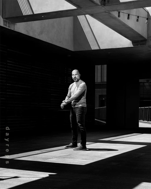 Monochrome portrait of a bald adult male in a long-sleeve shirt and jeans, standing in a sunlit architectural space with shadows.