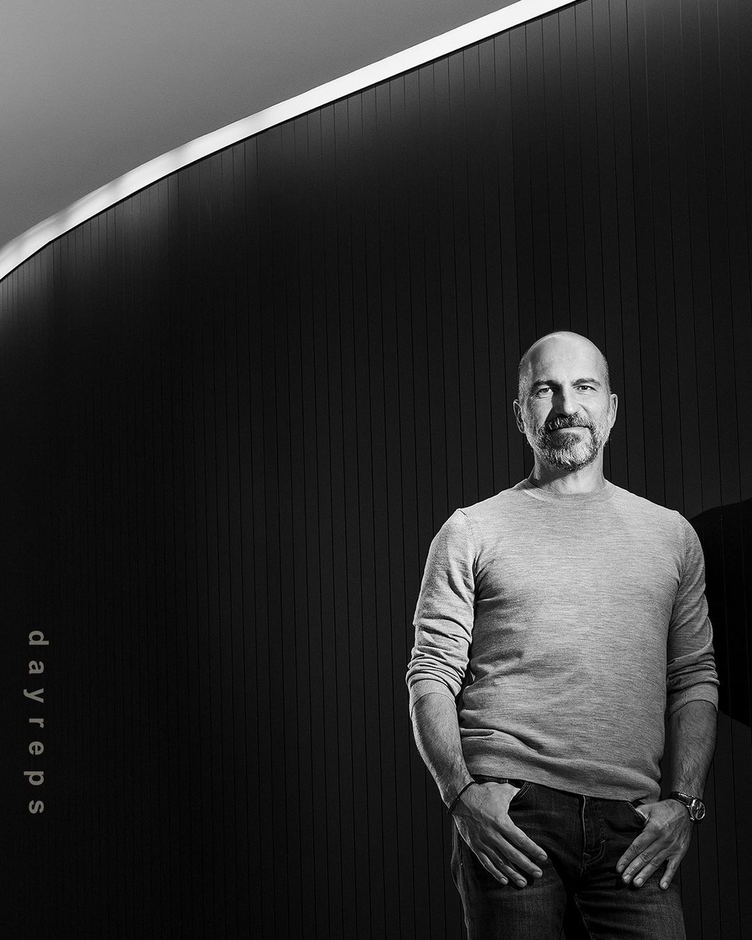 Portrait of a bald man with a beard wearing a knit sweater standing against a dark ribbed wall by Christie Hemm Klok