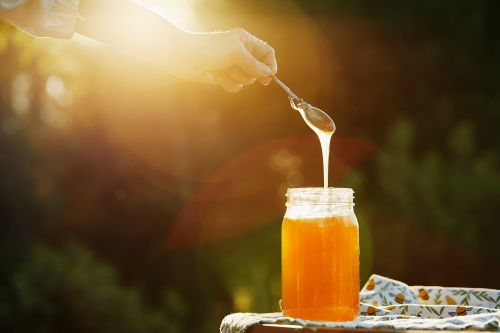 Golden honey pouring from a silver spoon into a clear glass jar, lit by bright sunshine against a blurry green natural background.