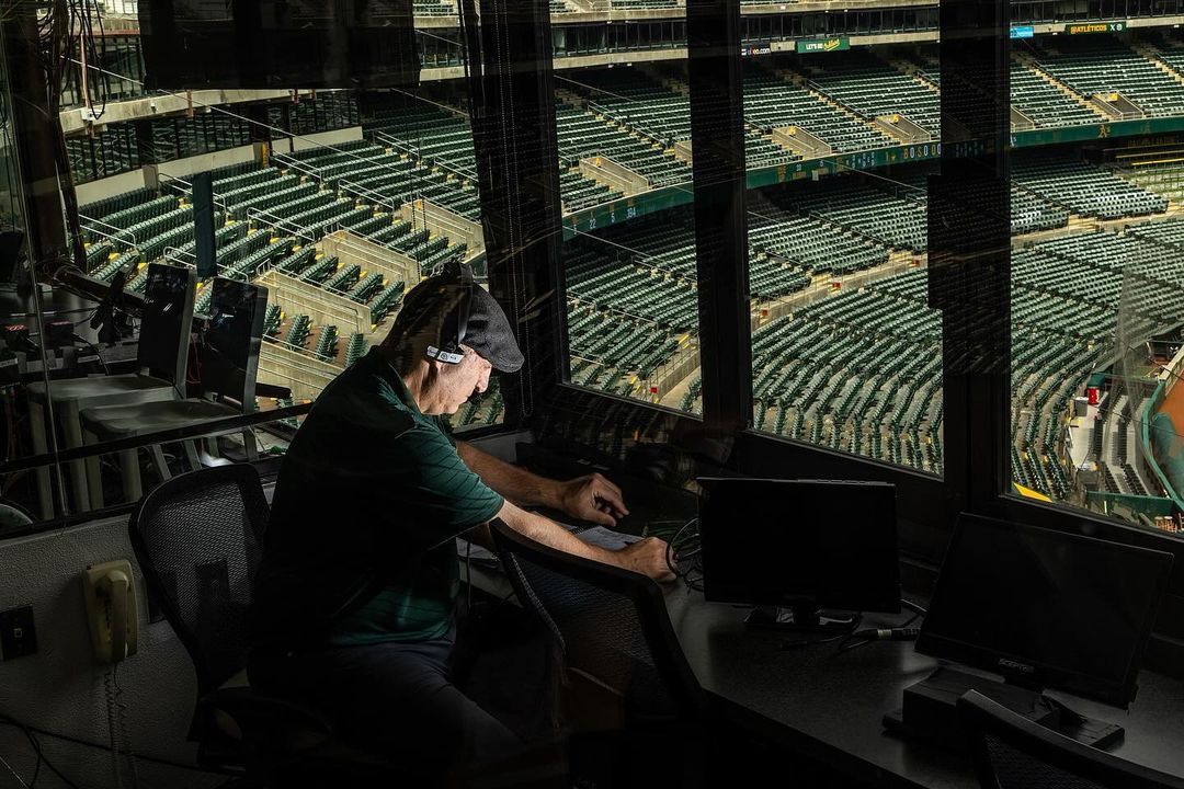 Man at work in a broadcast booth overlooking empty baseball stadium seating, with multiple monitors and production equipment visible.
