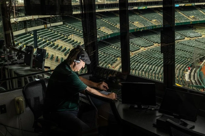 Man at work in a broadcast booth overlooking empty baseball stadium seating, with multiple monitors and production equipment visible.