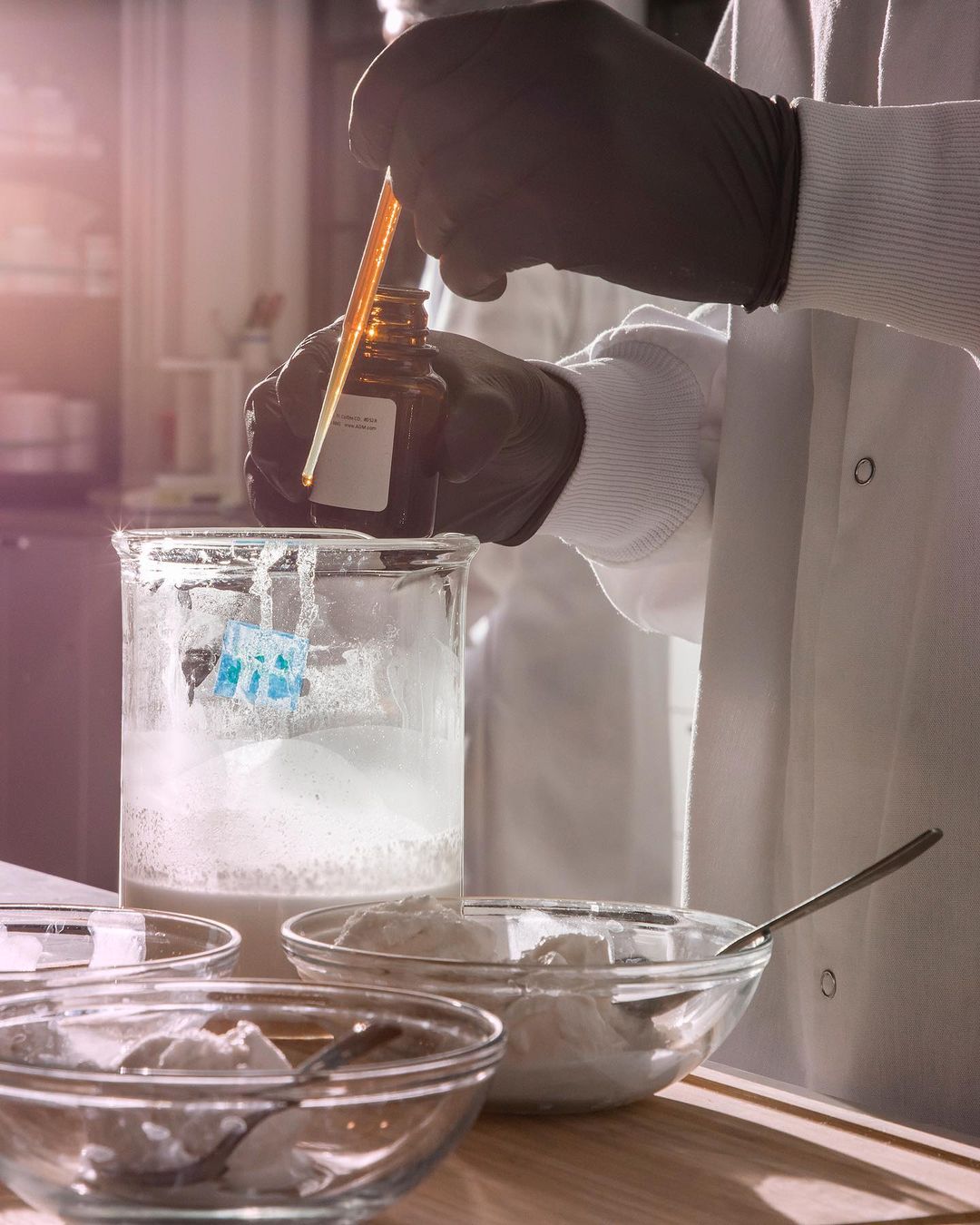 Person in lab coat holds dropper over beaker with white powder substance in laboratory setting; gloved hands, and glassware are visible.