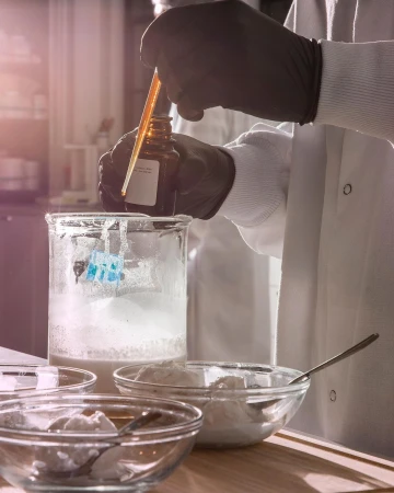 Person in lab coat holds dropper over beaker with white powder substance in laboratory setting; gloved hands, and glassware are visible.