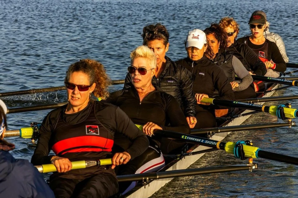 Eight women row a scull on a sunny waterway, oars dipped in the water, wearing matching team gear in a medium, eye-level outdoor shot.