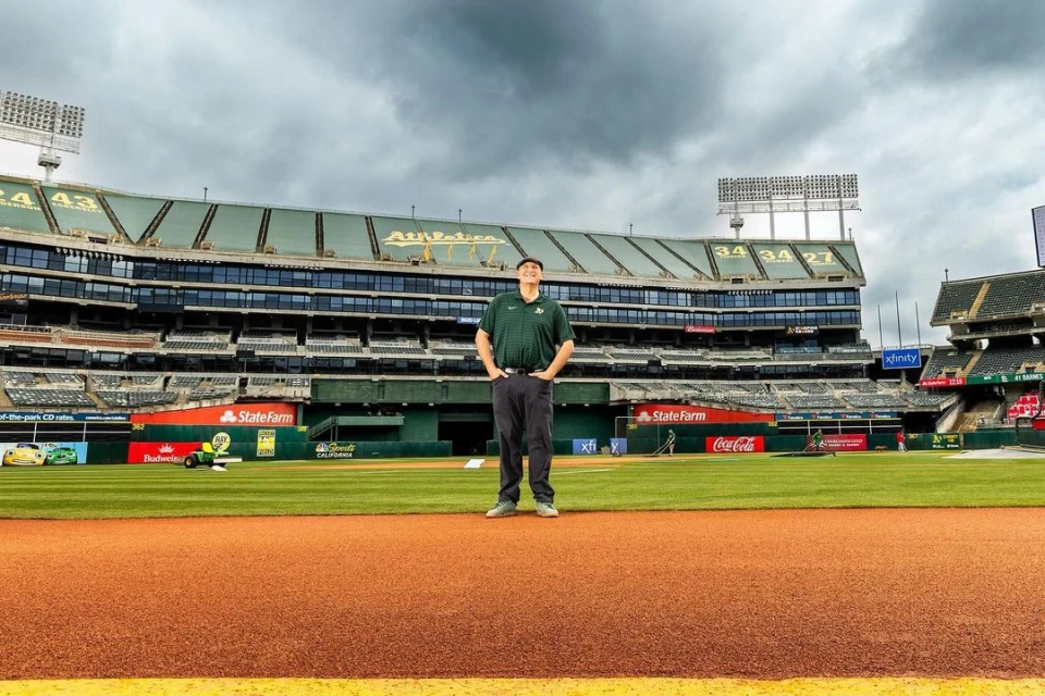 Man stands on a baseball field in front of stadium seating and architecture, under cloudy skies on a partly sunny day outdoors.