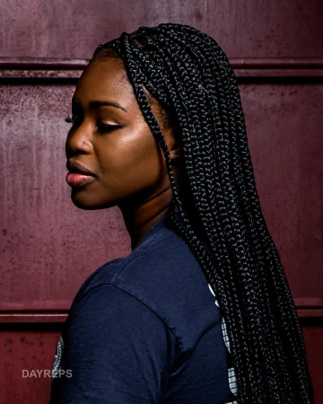 Profile portrait of a Black woman with long braided hair wearing a dark blue t-shirt in front of a rustic red metal wall.
