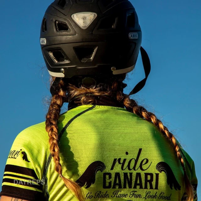 Back view of a cyclist with helmet and pigtails, wearing a neon green shirt with 'ride CANARI' logo against a clear blue sky.