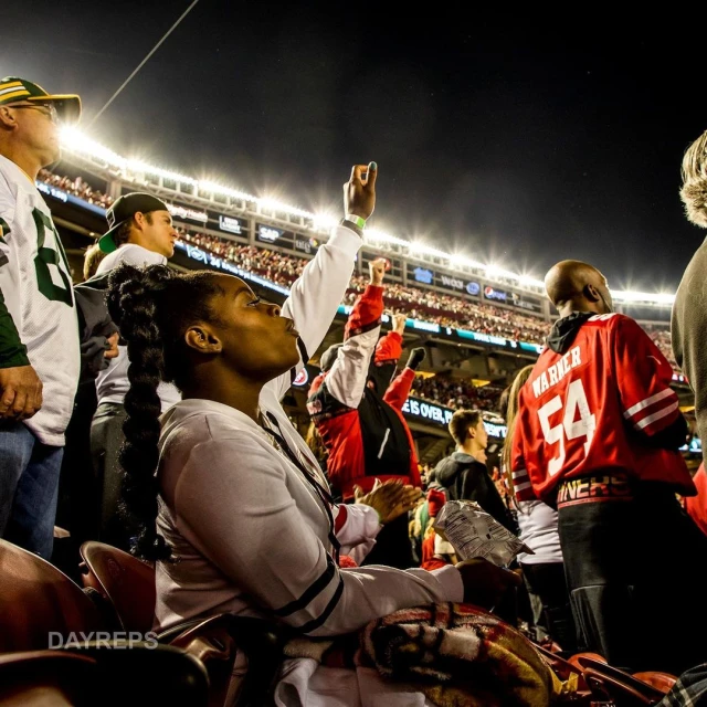 Spectators cheer at a football game at night, viewed from the stands, with stadium lights illuminating the crowd and the field.