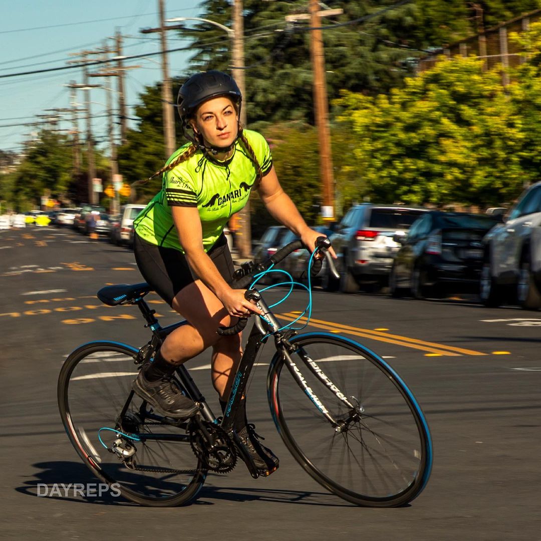 A woman rides a road bicycle down a city street, wearing a helmet and bright green cycling jersey, cars parked along the road.