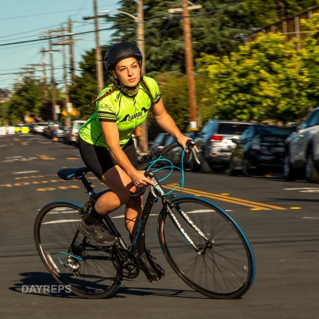 A woman rides a road bicycle down a city street, wearing a helmet and bright green cycling jersey, cars parked along the road.