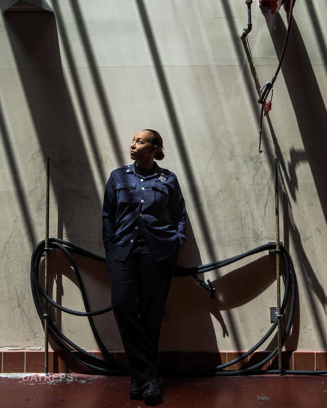 Adult woman in uniform poses against a wall with dramatic shadow patterns and nearby piping in an indoor, eye-level shot.
