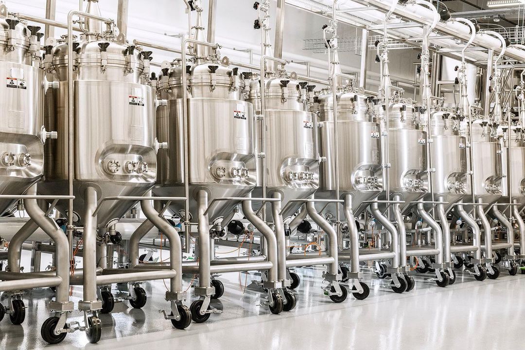 Stainless steel tanks on wheels dominate the interior of an industrial food processing facility, featuring metal piping and a clean, reflective floor.