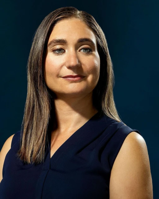 Close-up portrait of an adult woman with long hair, wearing a navy top, against a dark blue background, showcasing a professional headshot.