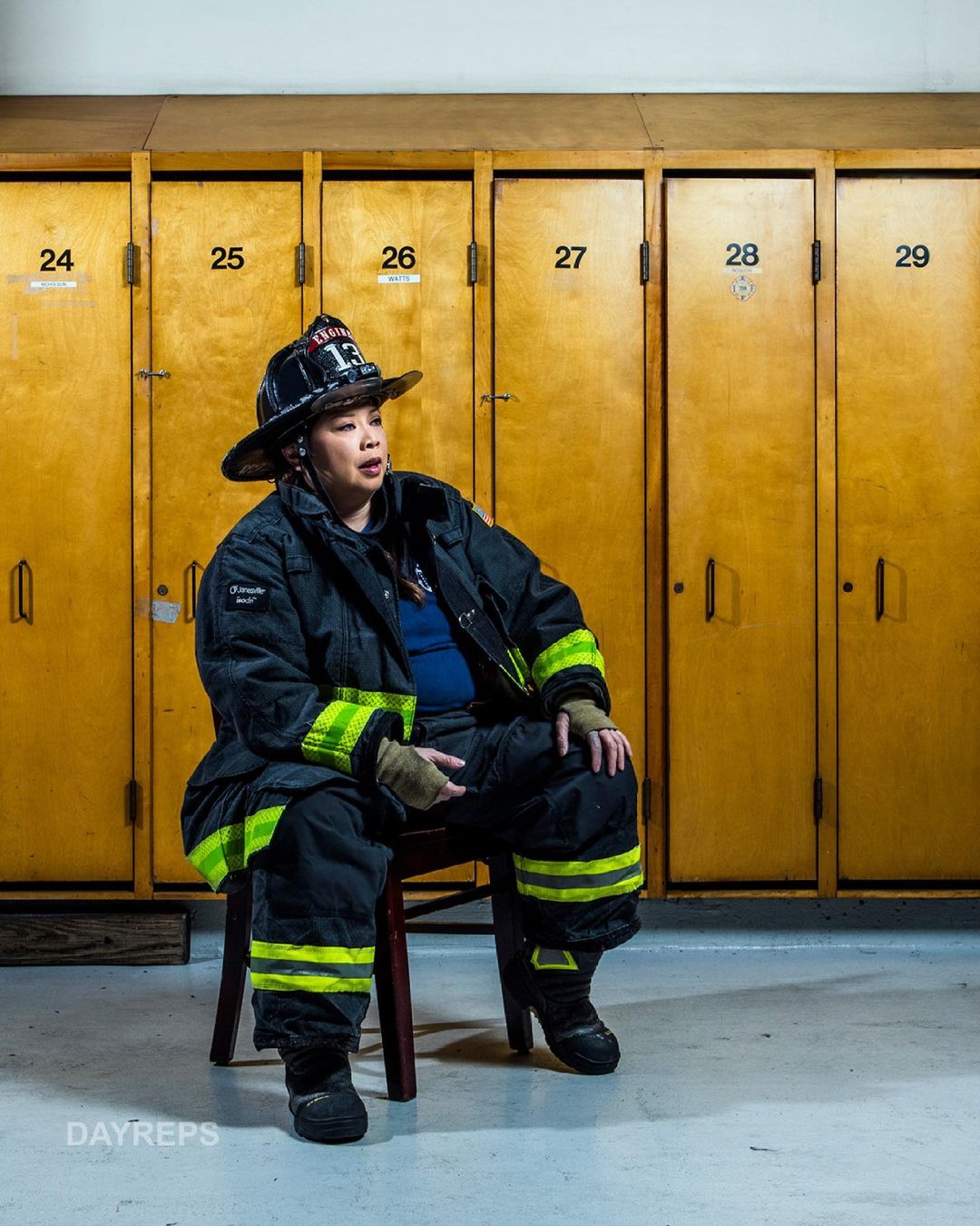 Female firefighter in uniform sits on a chair in front of lockers, wearing a helmet, jacket, and boots, indoors under bright lighting.