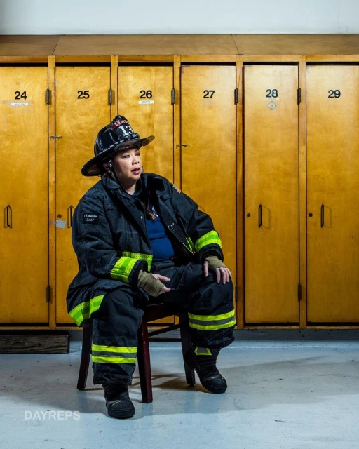 Female firefighter in uniform sits on a chair in front of lockers, wearing a helmet, jacket, and boots, indoors under bright lighting.