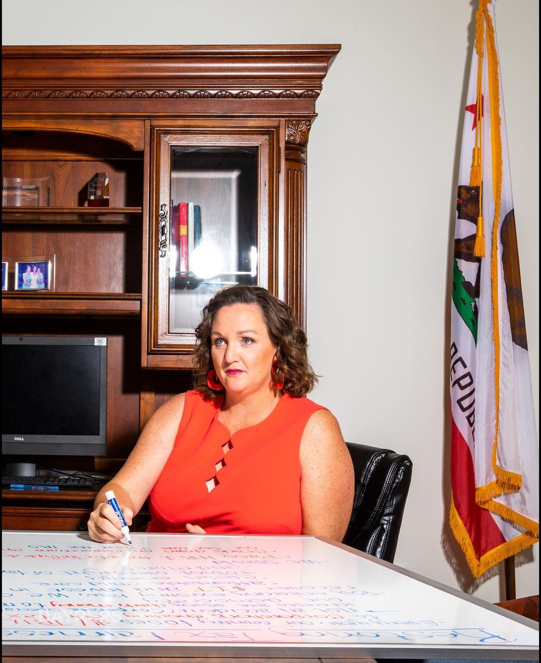 Woman in orange dress stands at a desk covered in writing, holding a marker, with a California flag and wooden cabinet behind.