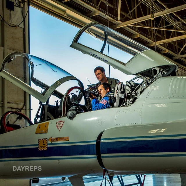 Two people inside the cockpit of a white jet airplane in an aircraft hangar, with the pilot holding a camera.