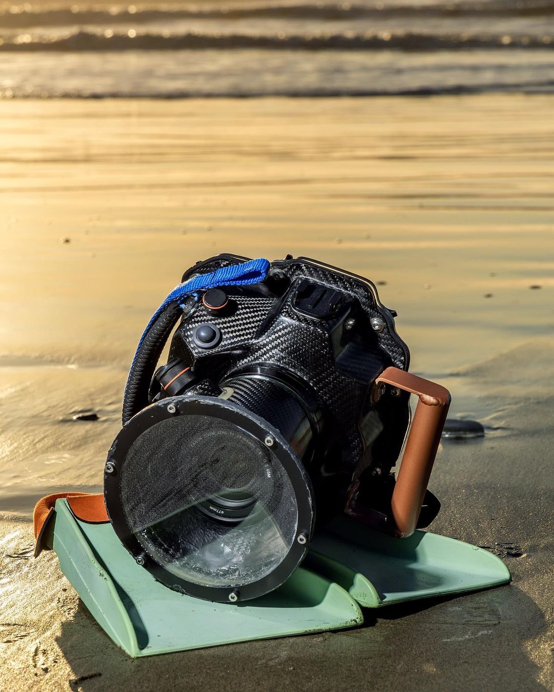 Underwater camera housing on sandy beach with ocean waves in background, showcasing water photography gear, and carbon fiber textures.