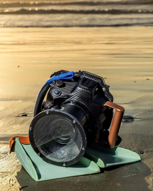 Underwater camera housing on sandy beach with ocean waves in background, showcasing water photography gear, and carbon fiber textures.