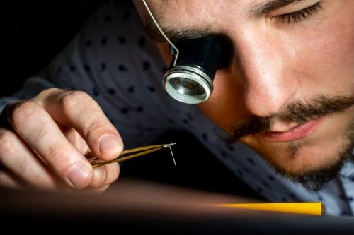 Close-up of a man using tweezers and a magnifying glass headpiece, focusing on detail, illuminated by a desk lamp. 