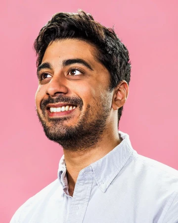 Portrait of a smiling South Asian man wearing a light blue buttoned shirt against a solid pink background.
