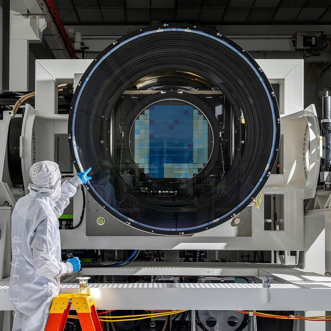 Scientist in clean suit points at a large blue wafer inside of a black lens of a white machine in a bright research lab.
